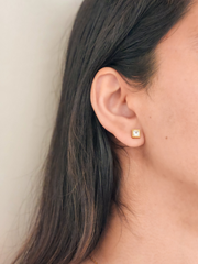 Close-up of an ear wearing a gold earring with a square gemstone on a neutral background