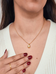 Woman wearing a gold necklace with a pendant, hand resting on chest.